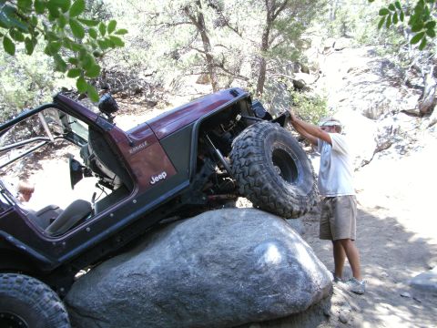 1995 Ford Bronco - A maroon Jeep Wrangler with the hood removed, roll cage, and oversized mud-terrain tires climbing a large granite boulder on a dusty desert…