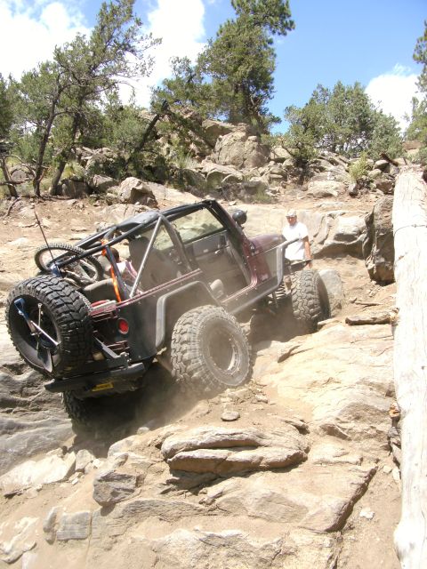 1995 Ford Bronco - Elevated three-quarter rear view of a topless maroon Jeep Wrangler with an exposed roll cage, rock sliders, tube rear bumper and swing-out…