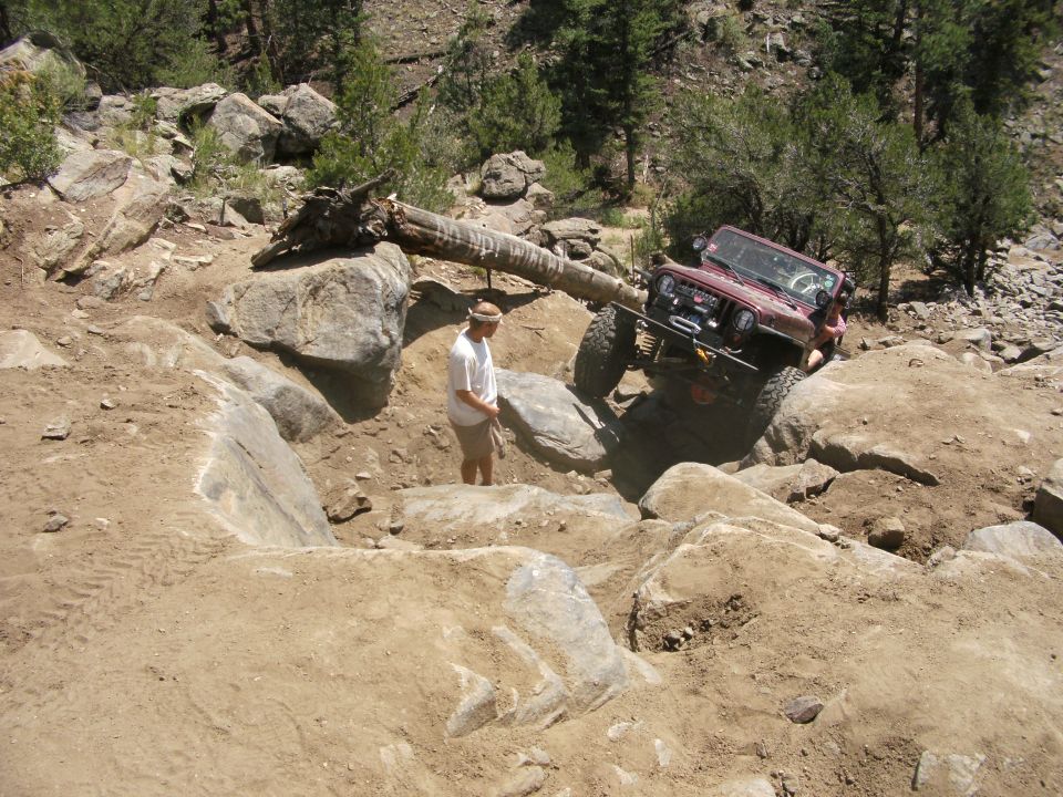 Red Jeep Wrangler navigating a difficult rocky obstacle on an off-road trail with a person standing nearby