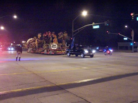 1990 Ford Bronco - A black pickup truck tows a parade float at night, with a police motorcycle escort.