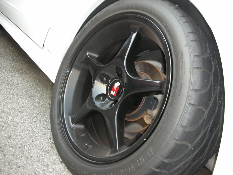 Close-up of a black five-spoke wheel with a red and white center cap on a car with a white body panel. Tire has visible text