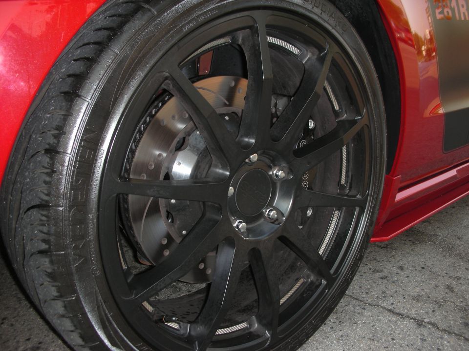Red car side view showing black multi-spoke wheels and drilled brake rotors