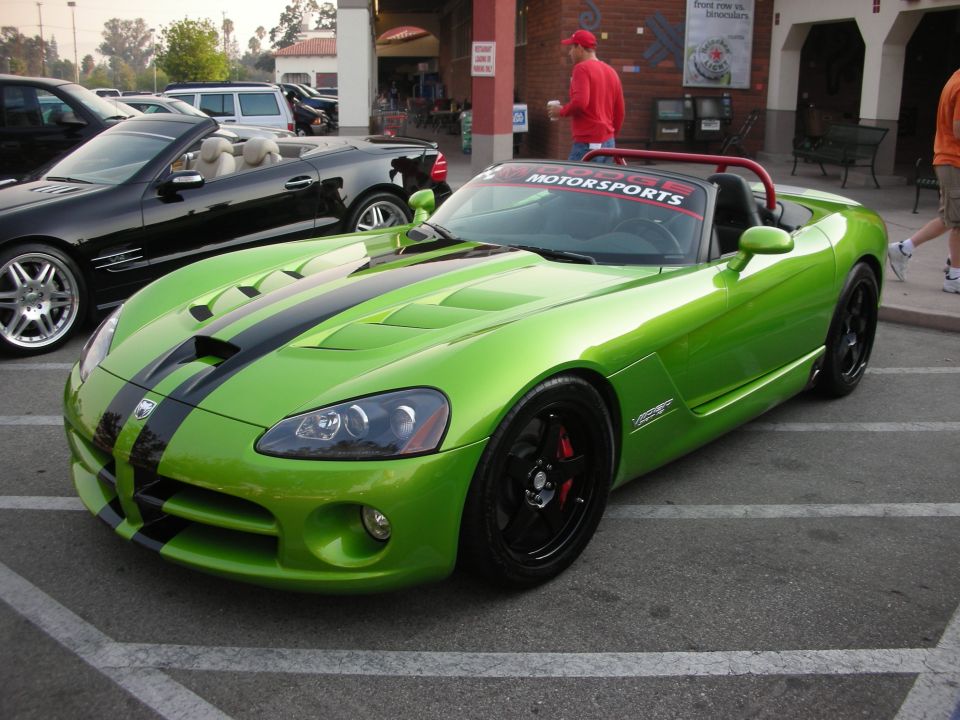 Lime green Dodge Viper convertible with black racing stripes and a 'MOTORSPORTS' banner on the windshield. Black wheels with red brake calipers visible. A black convertible is parked behind it