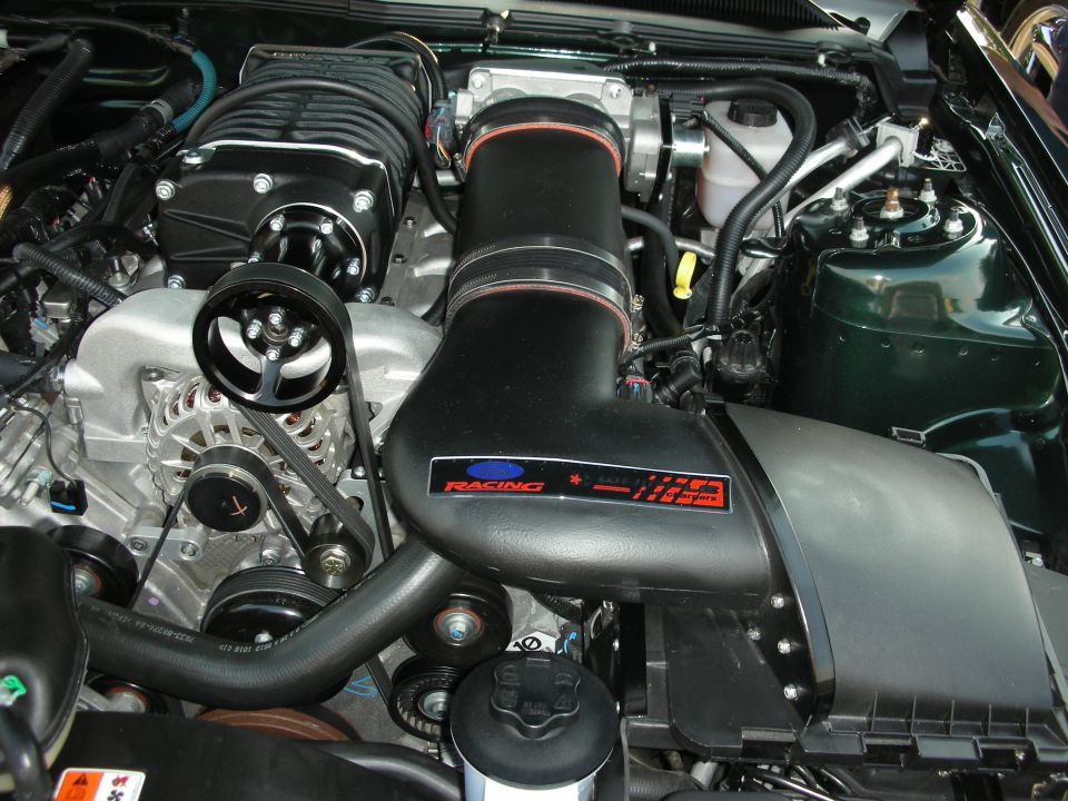 Engine bay with a black supercharger, intake manifold, and alternator. Some hoses and belts are visible