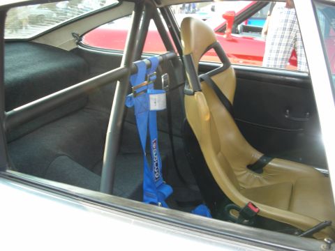 1998 Ford Crown Victoria - The interior of a car features a roll cage, a tan racing seat, and a blue Sparco harness, with other vehicles visible outside.