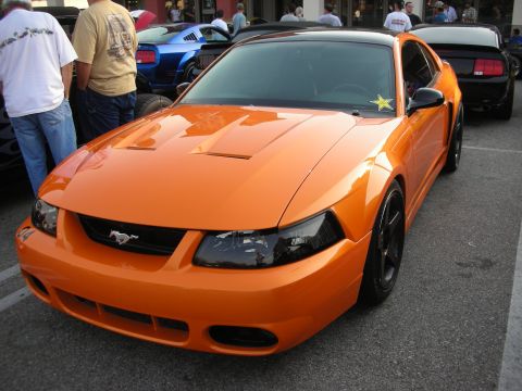 1998 Ford Crown Victoria - An orange Ford Mustang is parked at a car meet with other vehicles in the background.