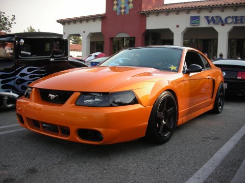 1998 Ford Crown Victoria - A bright orange Ford Mustang is parked in front of a building with a black roof and black wheels.