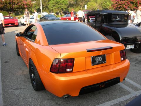 1998 Ford Crown Victoria - Rear view of a bright orange Ford Mustang with a black roof and tinted windows at a car gathering.