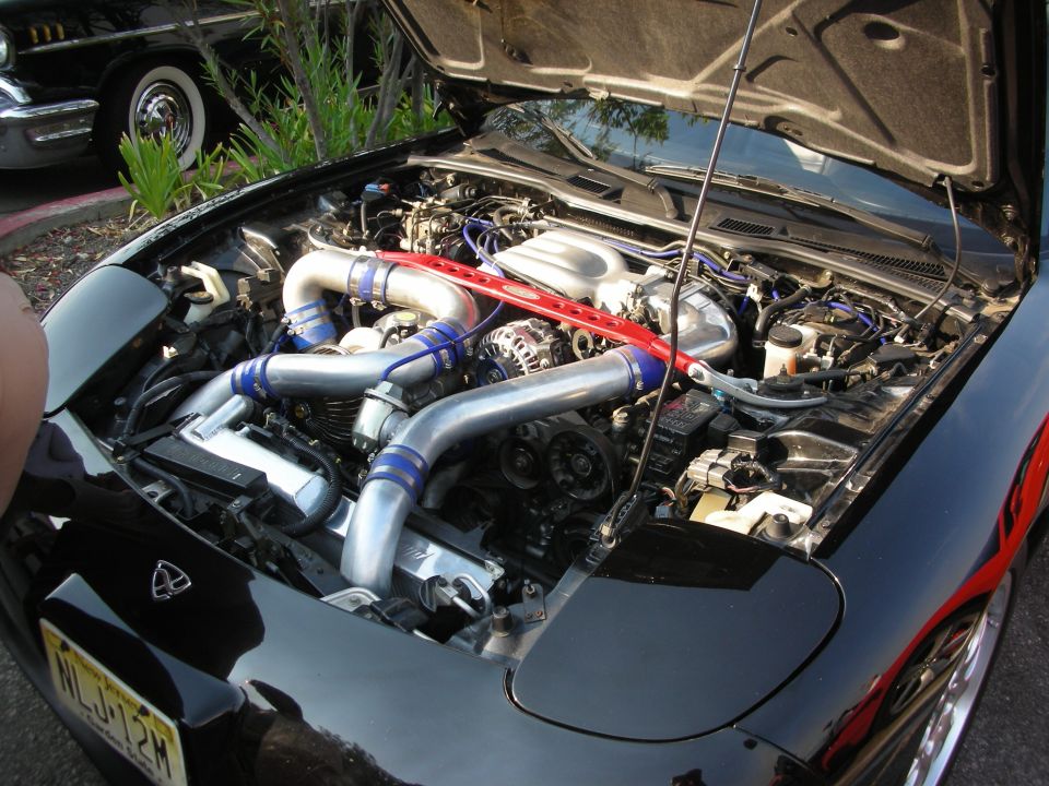 Black car engine bay with twin turbochargers and a red strut bar. A vintage black car is visible in the background