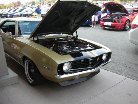 1998 Ford Crown Victoria - A gold 1969 Chevrolet Camaro is parked with its hood open, revealing the engine bay. Other vehicles are visible in the background.
