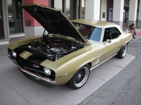 1998 Ford Crown Victoria - A gold 1969 Chevrolet Camaro Z/28 is parked with its hood open, revealing the engine bay and black racing stripes.