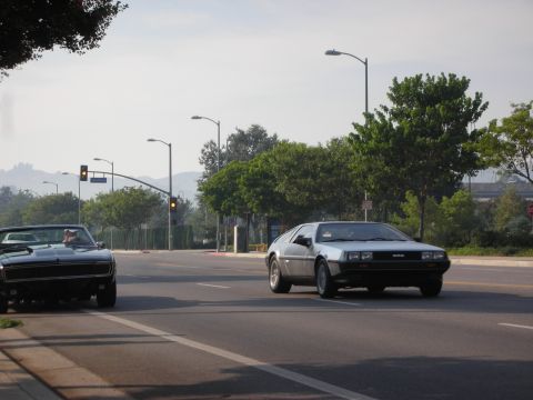 1998 Ford Crown Victoria - Two cars, a DeLorean and a muscle car, drive down a street on a sunny day.