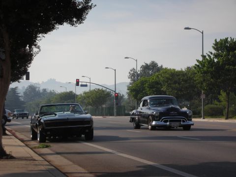 1998 Ford Crown Victoria - Two vintage cars cruise down a suburban street under a hazy sky.