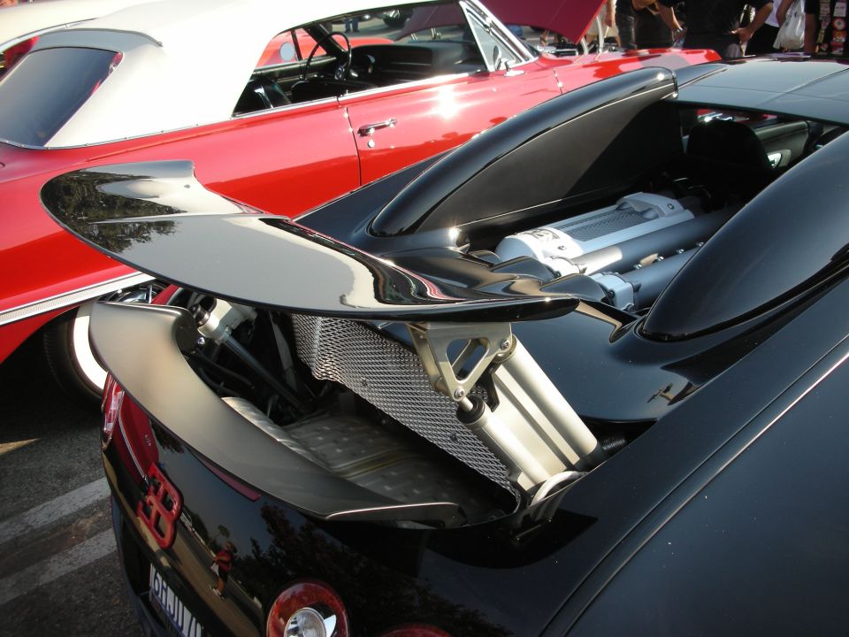 Black sports car's rear spoiler and exposed engine bay, with a red convertible in the background