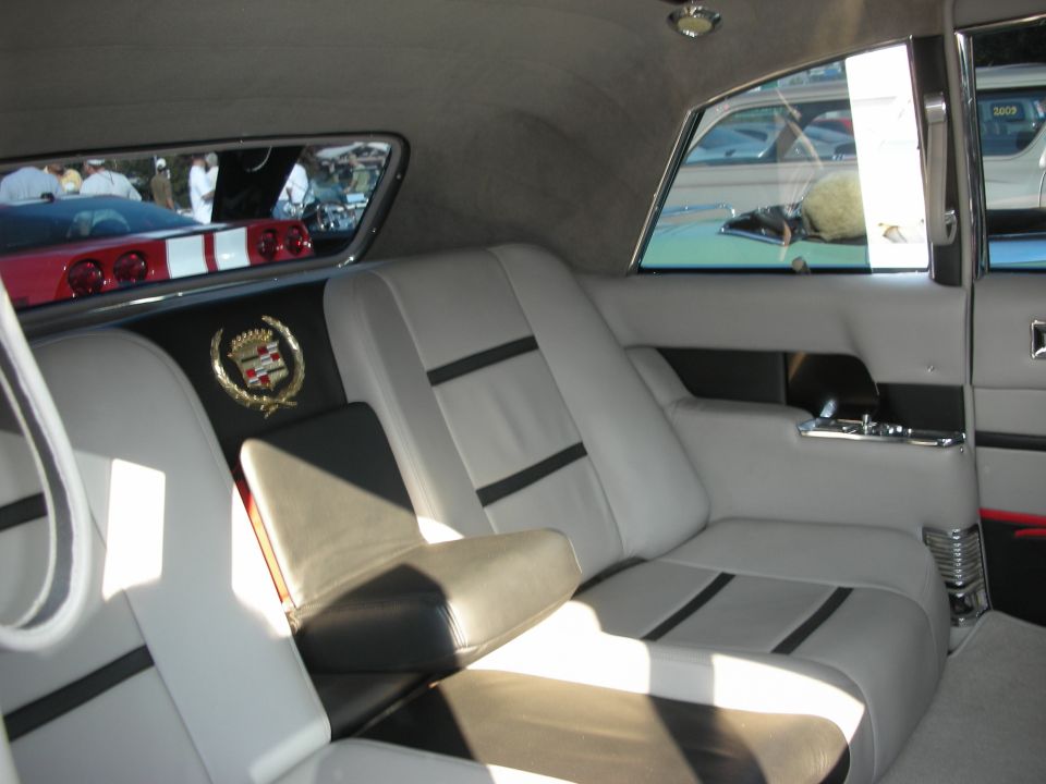 Interior of a car with white and black seats, a Cadillac emblem on the back panel, and reflections of other cars in the rear window
