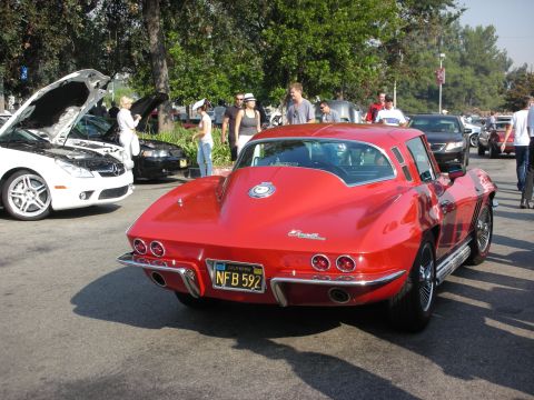 1998 Ford Crown Victoria - A red 1963-1967 Chevrolet Corvette Sting Ray coupe is parked at a car meet with other vehicles and people around.