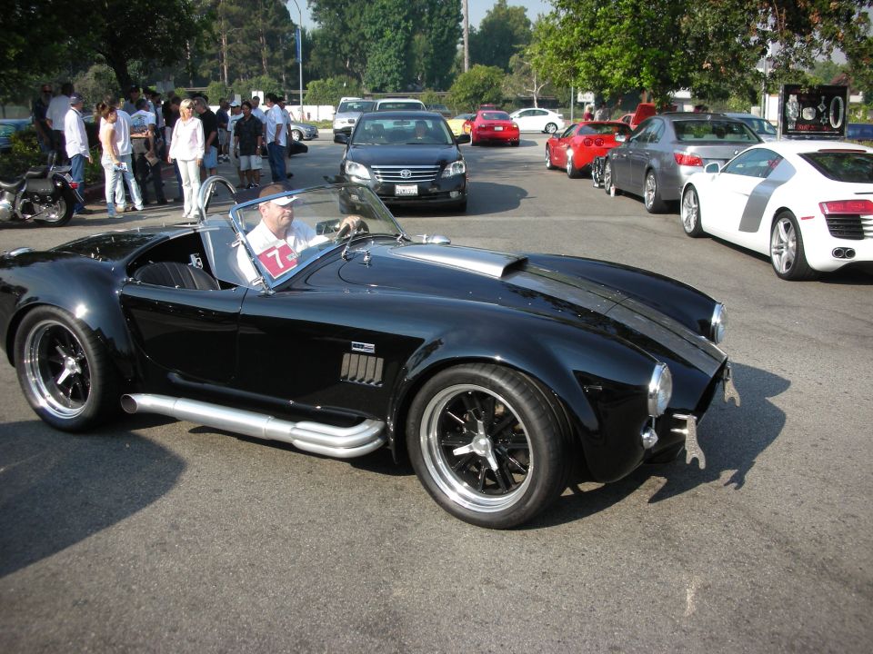 Black AC Cobra replica, driver wearing white shirt and hat, side pipes, silver hood scoop