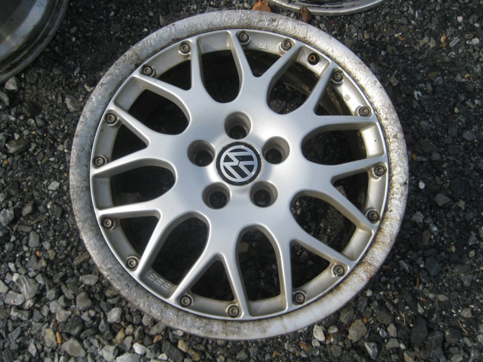 Silver Volkswagen alloy wheel with a BBS logo and a circular center cap with the Volkswagen emblem, resting on gravel