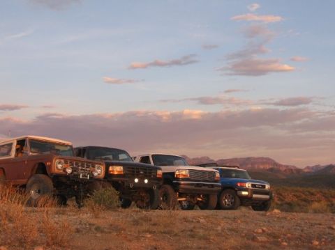 1996 Ford Bronco - Four off-road vehicles, including a 1992 Ford Bronco and a Toyota FJ Cruiser, are lined up on a desert hillside at sunset.