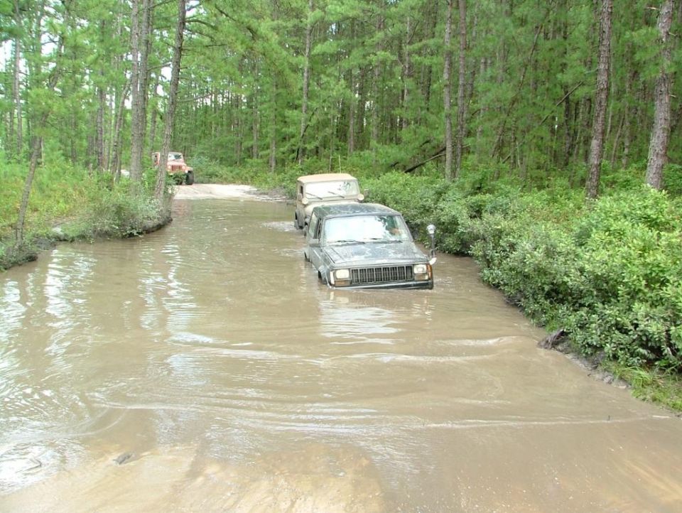 Gray Jeep Cherokee driving through muddy water, front-left quarter view. Other vehicles visible in background