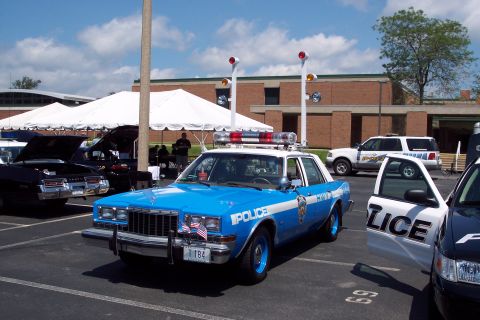 2001 Ford Crown Victoria - A blue and white police car with a light bar and American flags on the front is parked in a lot with other vehicles.