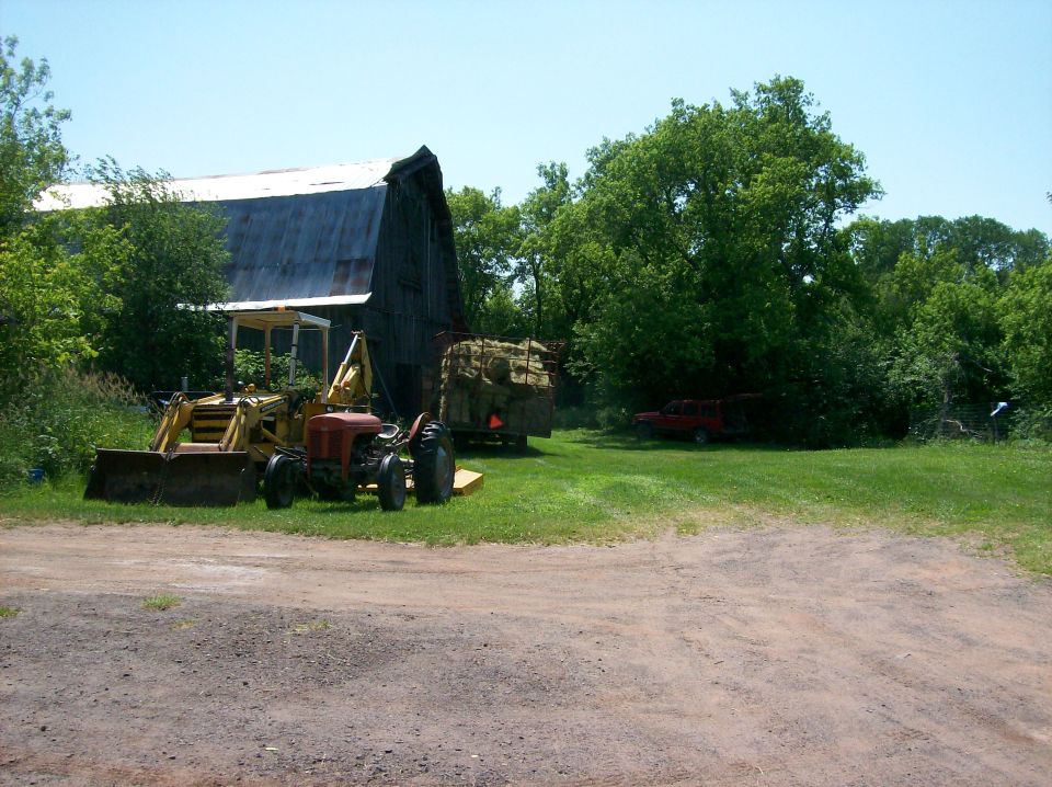 Tractor with front loader and trailer of hay bales next to a barn, with a red SUV in the background