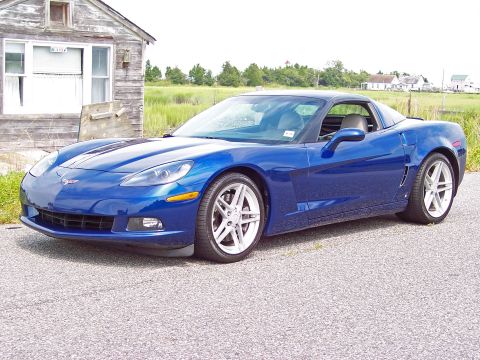 2006 Chevrolet Corvette - A blue Chevrolet Corvette is parked on an asphalt surface in front of a rustic wooden structure with a grassy field in the background.