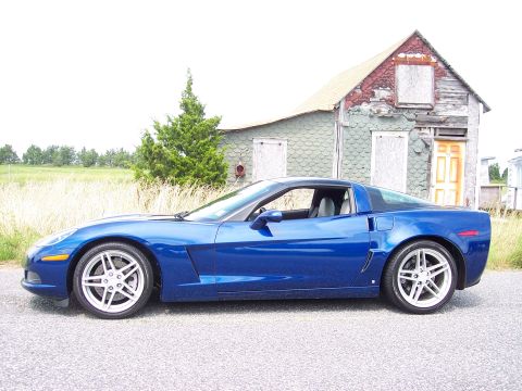 2006 Chevrolet Corvette - A blue sports car is parked on a gravel road in front of a dilapidated house with boarded-up windows.