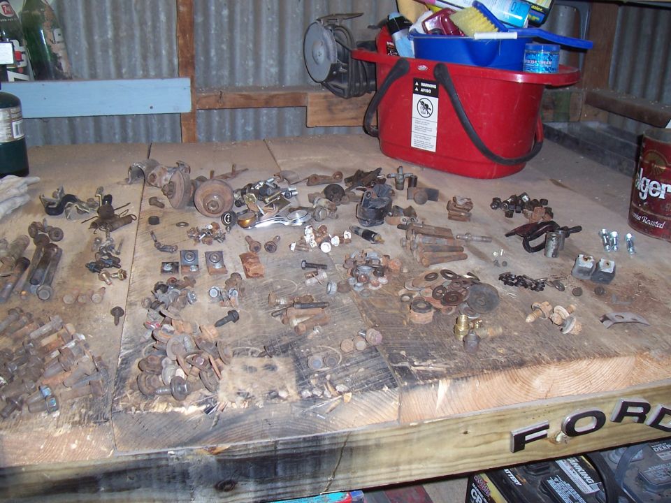 Table covered with various rusty car parts, bolts, nuts, and washers. A red bucket with cleaning supplies is in the background