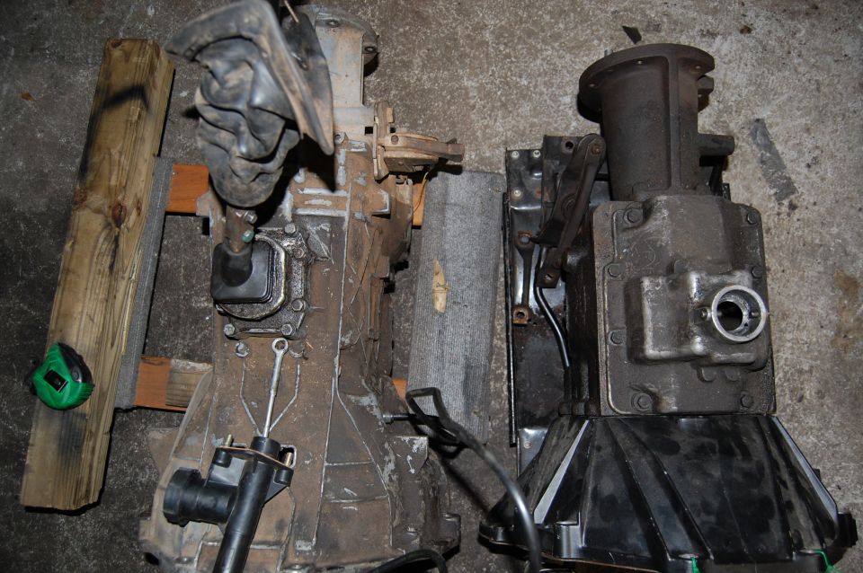 Two vehicle transmissions, one with a shifter boot, sit on a concrete floor next to wooden blocks