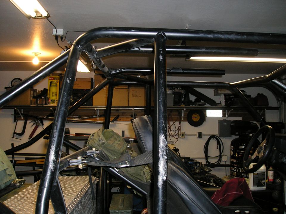 Interior view of a vehicle with a prominent black roll cage, seats, and a steering wheel. Shelving with tools and boxes is visible in the background