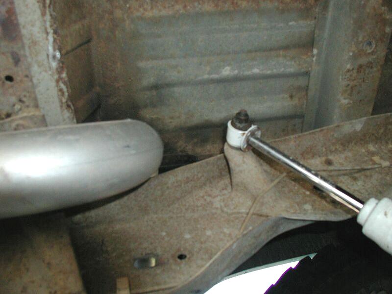 Underside of vehicle showing exhaust component, rusty floor pan, and tool loosening a nut