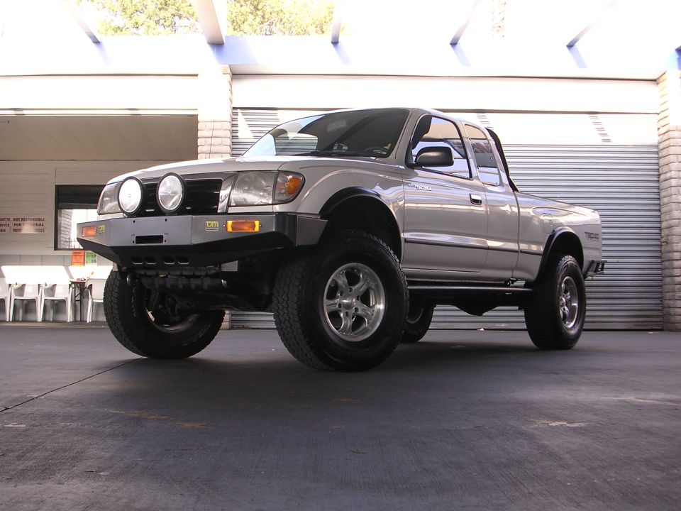 Silver Toyota pickup truck with aftermarket bumper and off-road tires, viewed from the front three-quarter angle