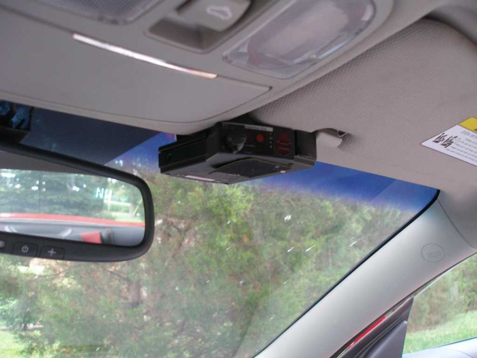 Black radar detector mounted to a gray headliner next to the rearview mirror