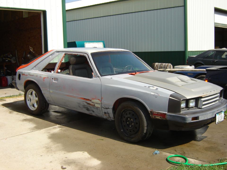 Primer-grey Mercury Capri hatchback, front 3/4 view, parked by a shed