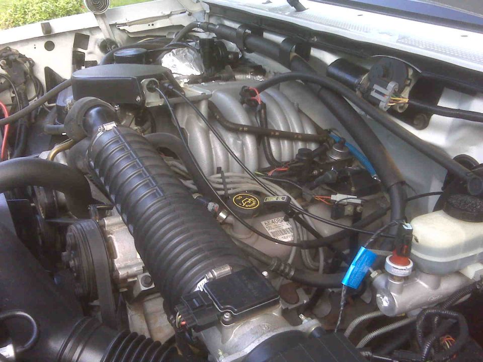 Close-up of a vehicle's engine bay with various hoses, wires, and engine components visible. A Ford logo is visible on the engine