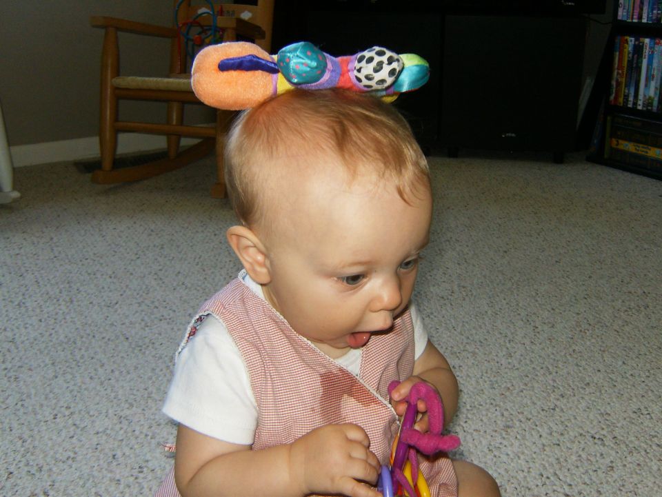 Baby wearing a colorful caterpillar toy on its head, holding a toy in its hands