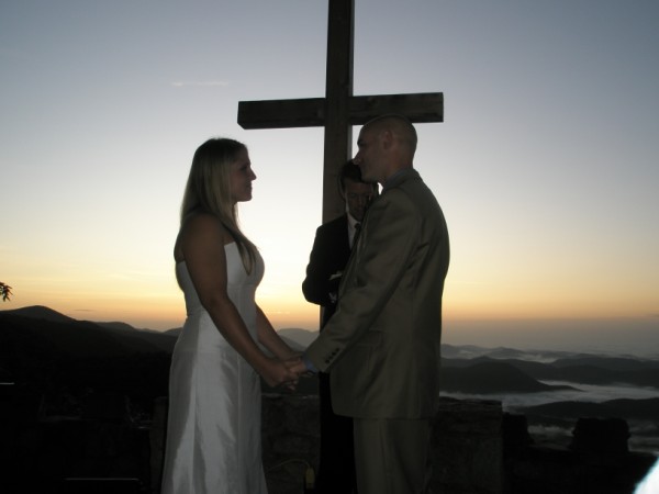 Bride and groom hold hands at sunset beneath a wooden cross on a mountain overlook