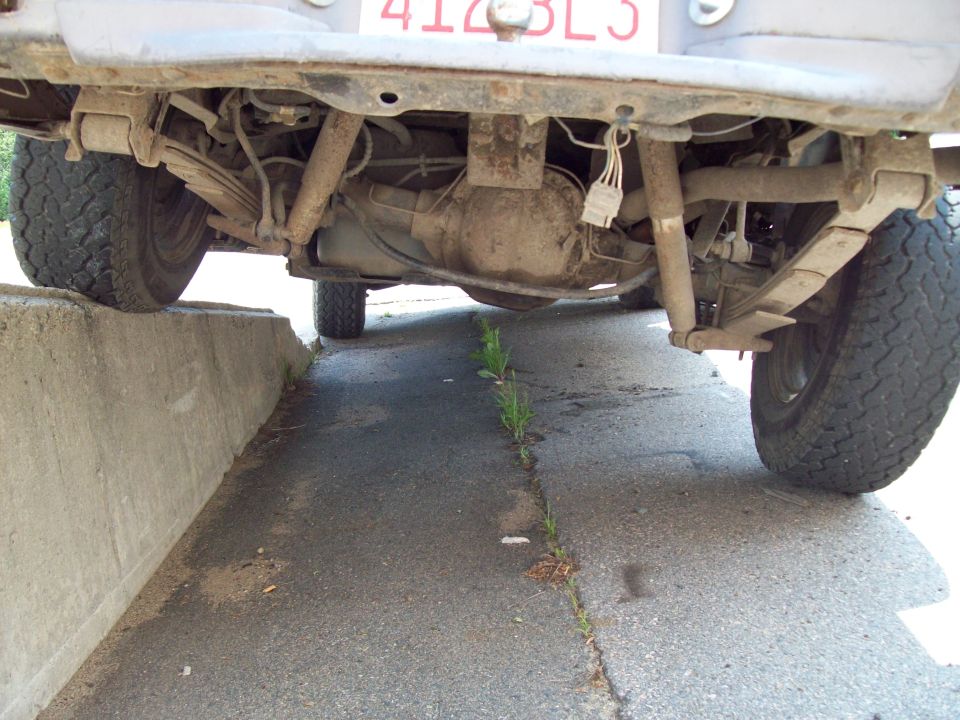 Underside rear view of an SUV showing rear axle, leaf springs and spare tire, one wheel up on a concrete curb.