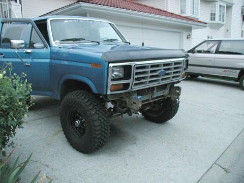 1985 Ford Bronco - A blue pickup truck is parked in a driveway with its front bumper removed, revealing the chassis and suspension components.