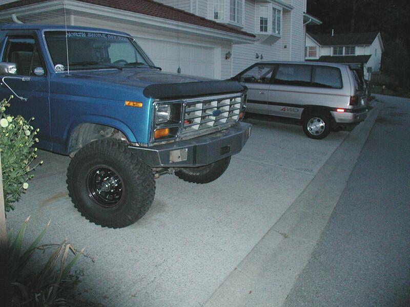 Blue lifted Ford Bronco with black wheels and gray bumper parked in driveway near silver minivan