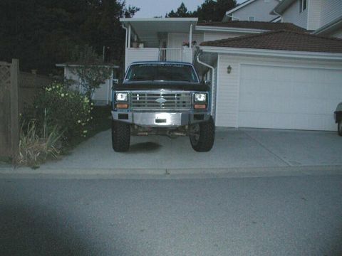 1985 Ford Bronco - A Ford Bronco is parked in front of a garage, showing its front grille and bumper.