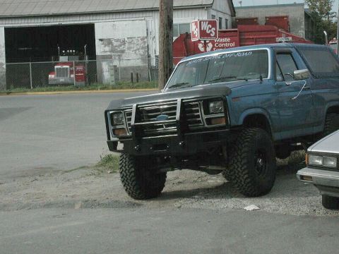 1985 Ford Bronco - A blue Ford Bronco is parked on a gravel lot, showing its front end and modified bumper.