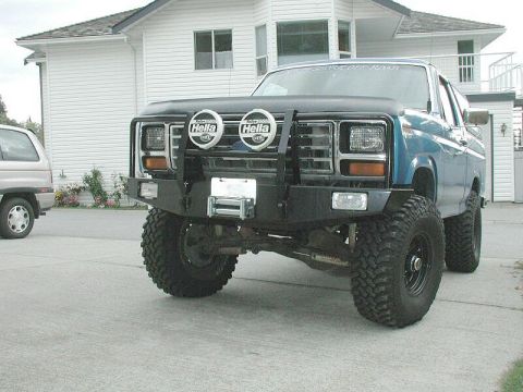 1985 Ford Bronco - This blue SUV features a aftermarket bumper with a winch and Hella lights, parked next to another vehicle.