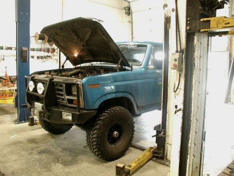 1985 Ford Bronco - Blue pickup truck on a lift in a garage, hood open, showing engine bay.