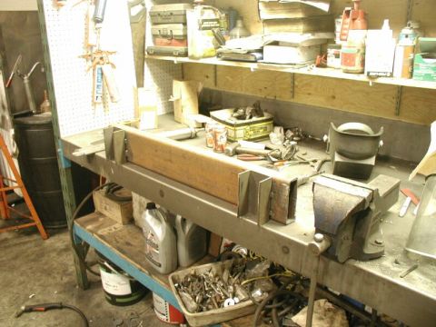 1985 Ford Bronco - A cluttered workbench holds various tools, oil containers, and metal parts, suggesting a workshop environment.