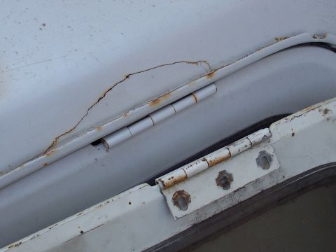 1970 Jeep Jeepster - Close-up of a rusted hinge on a white vehicle part, showing signs of wear and cracking.