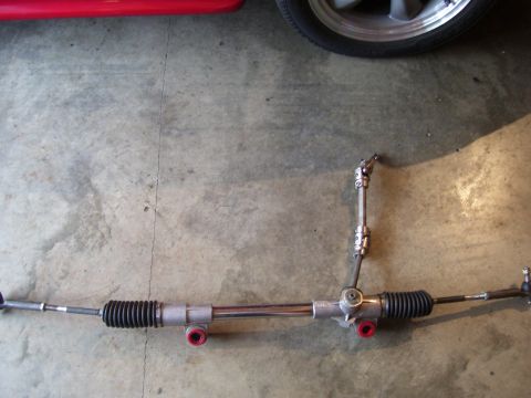 1990 Ford Mustang - A steering rack is shown on a concrete surface, with a portion of a red vehicle and a wheel in the background.