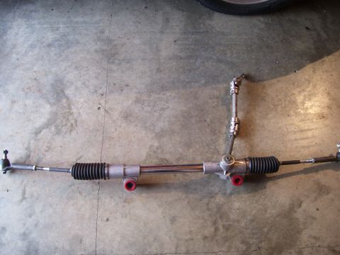 1990 Ford Mustang - A steering rack and pinion assembly is shown, red bushings and tie rod ends, laid out on a concrete floor.