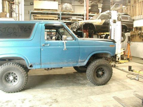 1985 Ford Bronco - A blue SUV is parked in a garage, with another vehicle lifted on a hoist in the background.